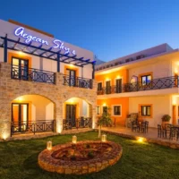 Aegean Sky Hotel & Suites, Malia—dusk scene with lit balconies, stone arches and a neat courtyard ready for your night ahead.