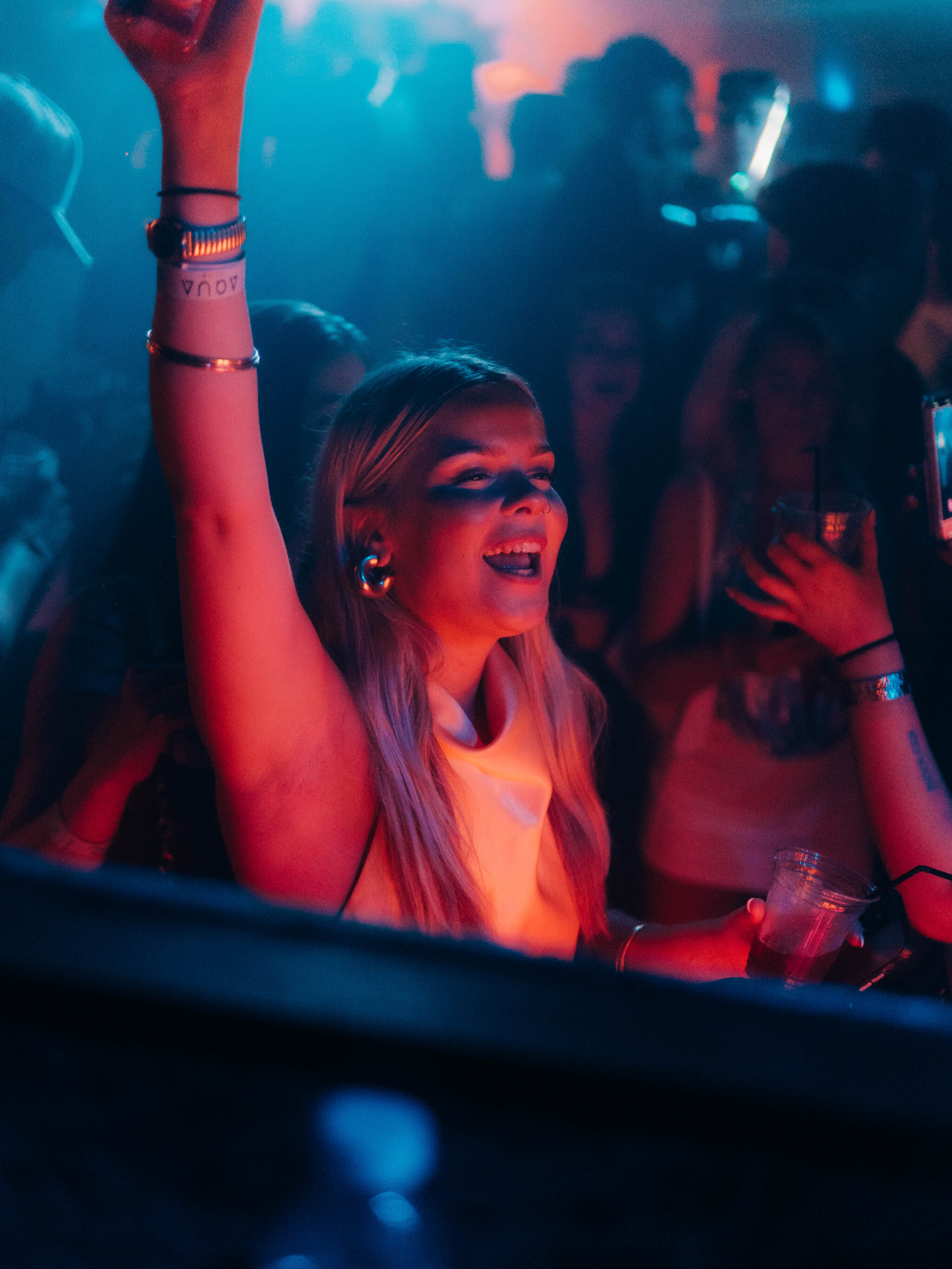 Woman with a drink in hand dancing with her arm raised under vibrant lights at the Lovejuice Residency event in Ayia Napa.