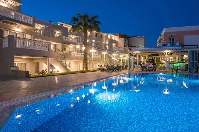 Sarpidon Apartments Malia lit up at dusk, showing the pool, balconies, palm trees and a relaxed holiday atmosphere.