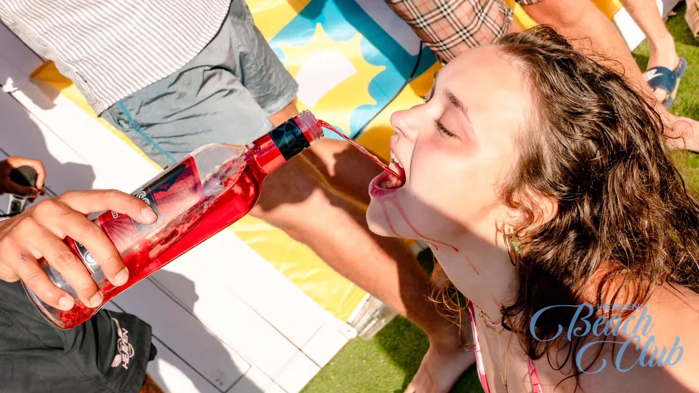 Girl at Albufeira Beach Club pool party drinking directly from a bottle of red alcohol in the sun.