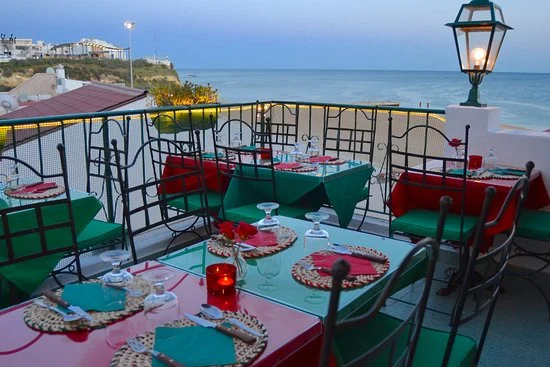 Scenic outdoor seating at Ricardo's Pizza Restaurant Albufeira, with red and green tables overlooking the sea at dusk.