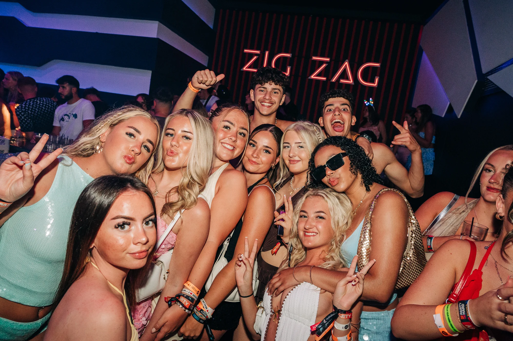 Group of girls and lads partying at Zig Zag Club in Malia, posing for a photo mid-night out with drinks, lights and big summer energy.