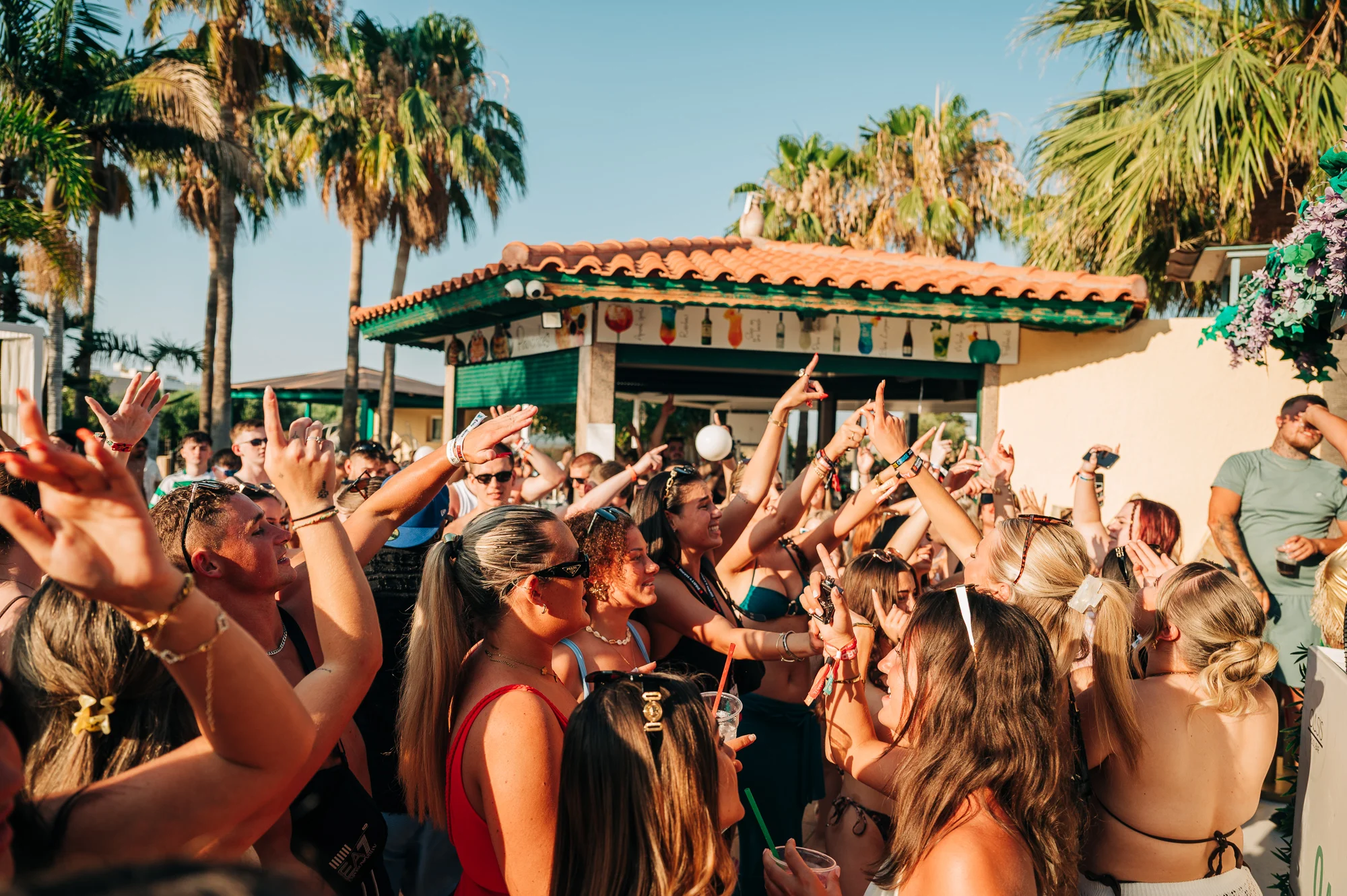 Crowds going wild under the sun at a Malia pool party, hands in the air and drinks flowing to the summer beats.