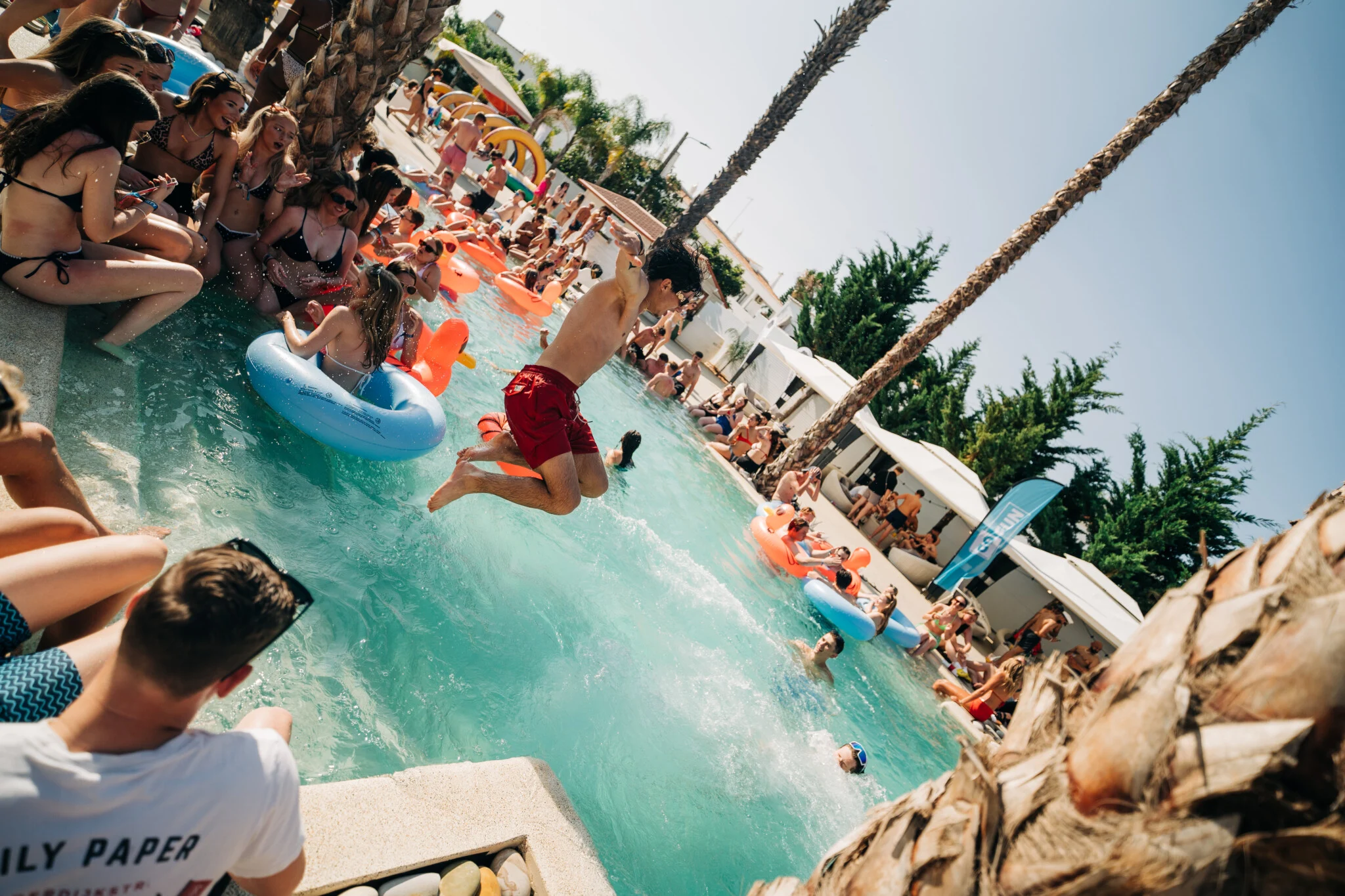 Pool party in Albufeira with people jumping in, inflatables floating and the sun shining over a buzzing crowd.