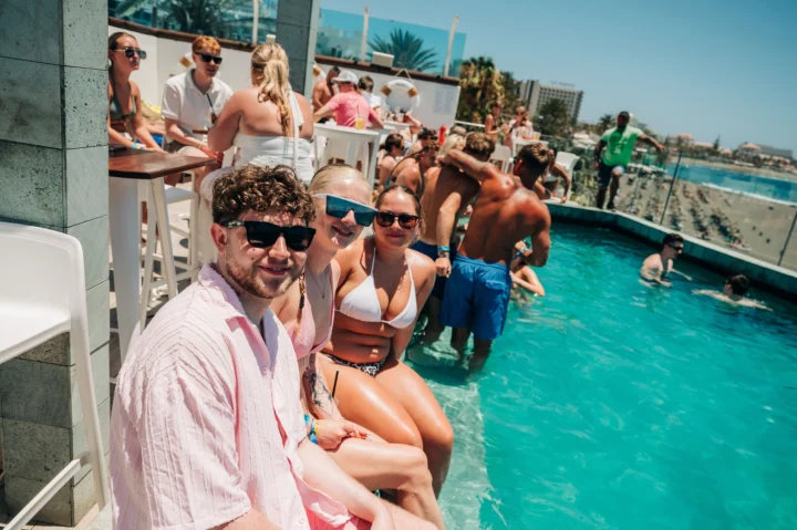 Girls and lads chilling poolside at a Tenerife day party, drinks flowing, sunshine blazing and beach views behind