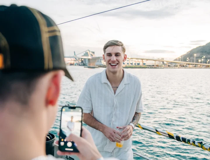 Friends capturing a sunset boat moment in Barcelona as a guy laughs with a drink, setting the vibe for a lively night.