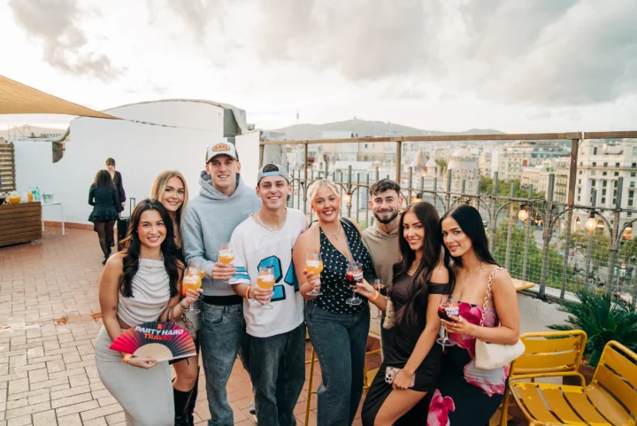 Friends raising drinks at a Barcelona rooftop party, sunset warm-up vibes, city views and the night about to kick off