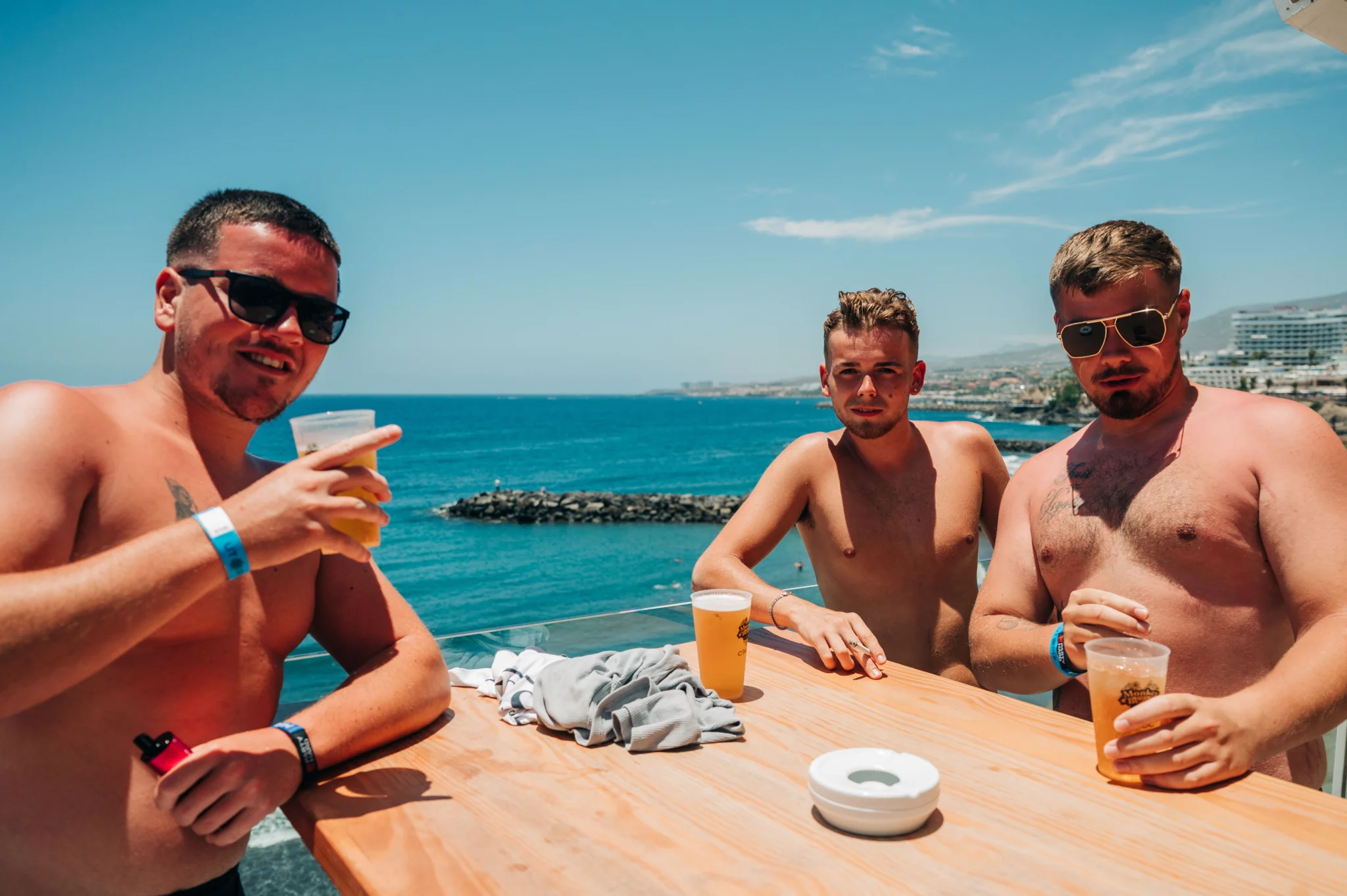 Lads drinking beers at a Tenerife beach bar, sun out, sea behind, chilled pre-party vibes before the night