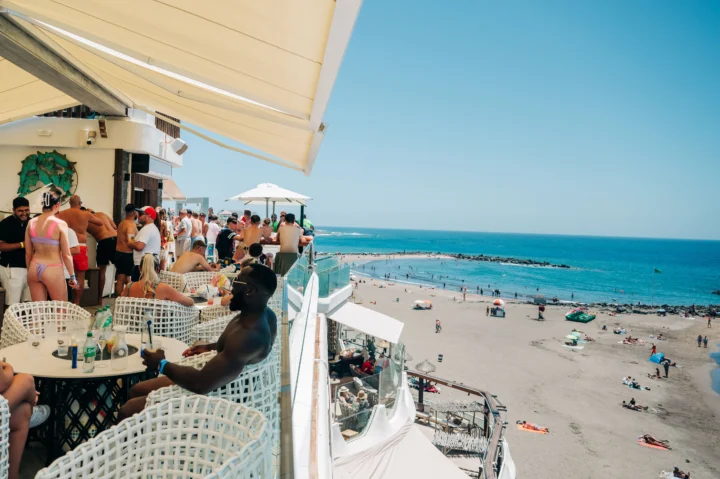 Beachfront bar party in Tenerife with lads and girls drinking, sun blazing, music pumping above the sand and sea