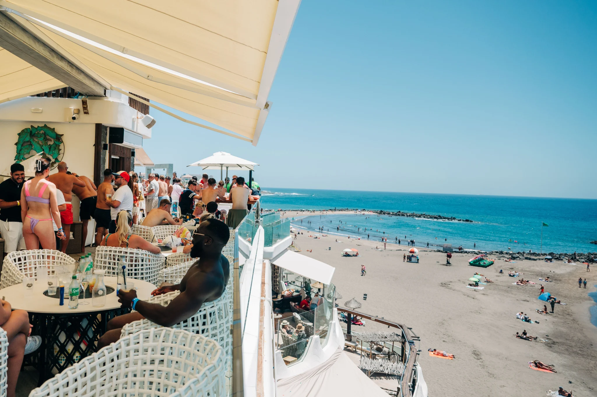 Beachfront bar party in Tenerife with lads and girls drinking, sun blazing, music pumping above the sand and sea