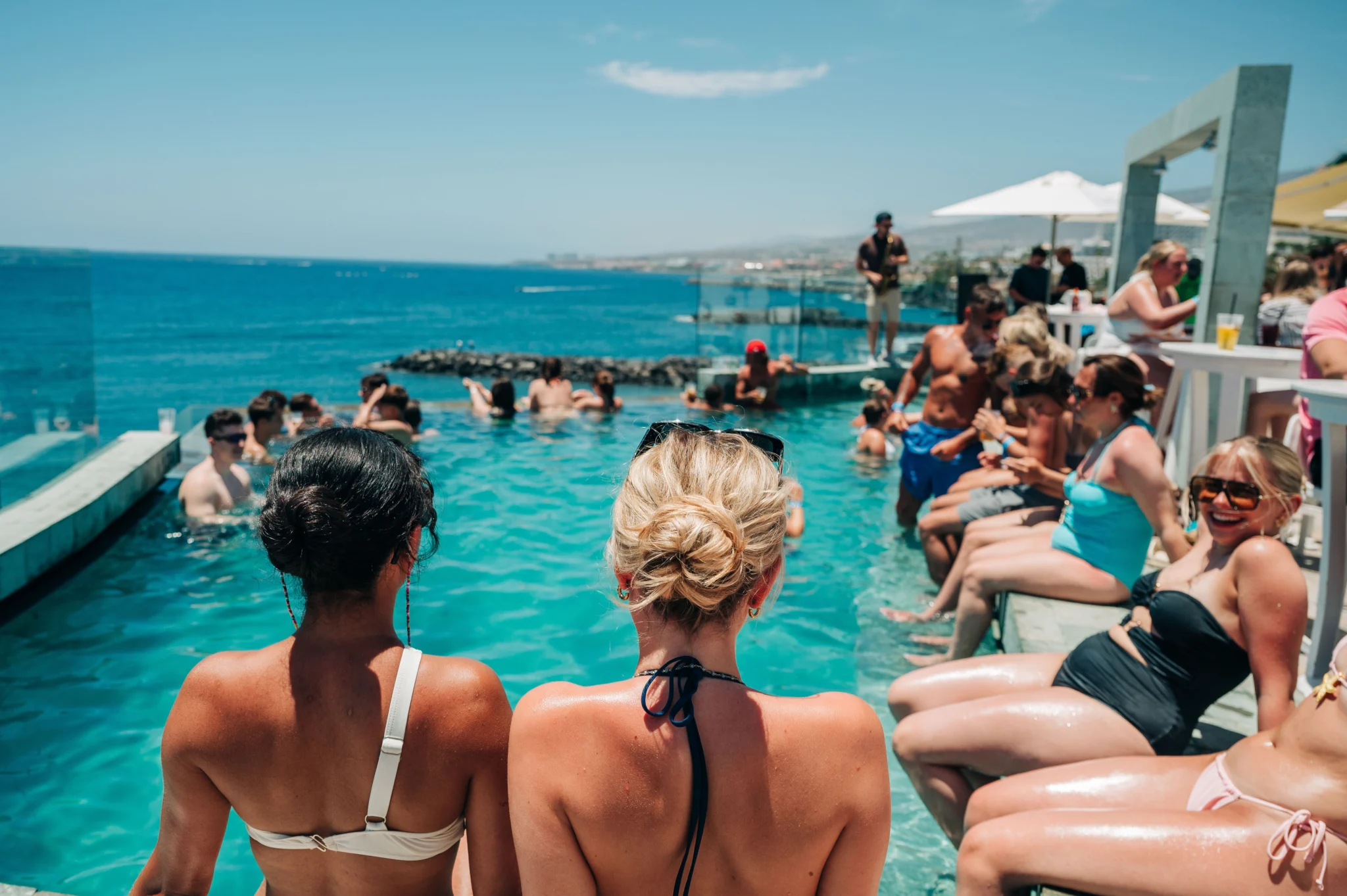 Girls and lads lined up poolside at a Tenerife day party, feet in the water with ocean views and a buzzing crowd
