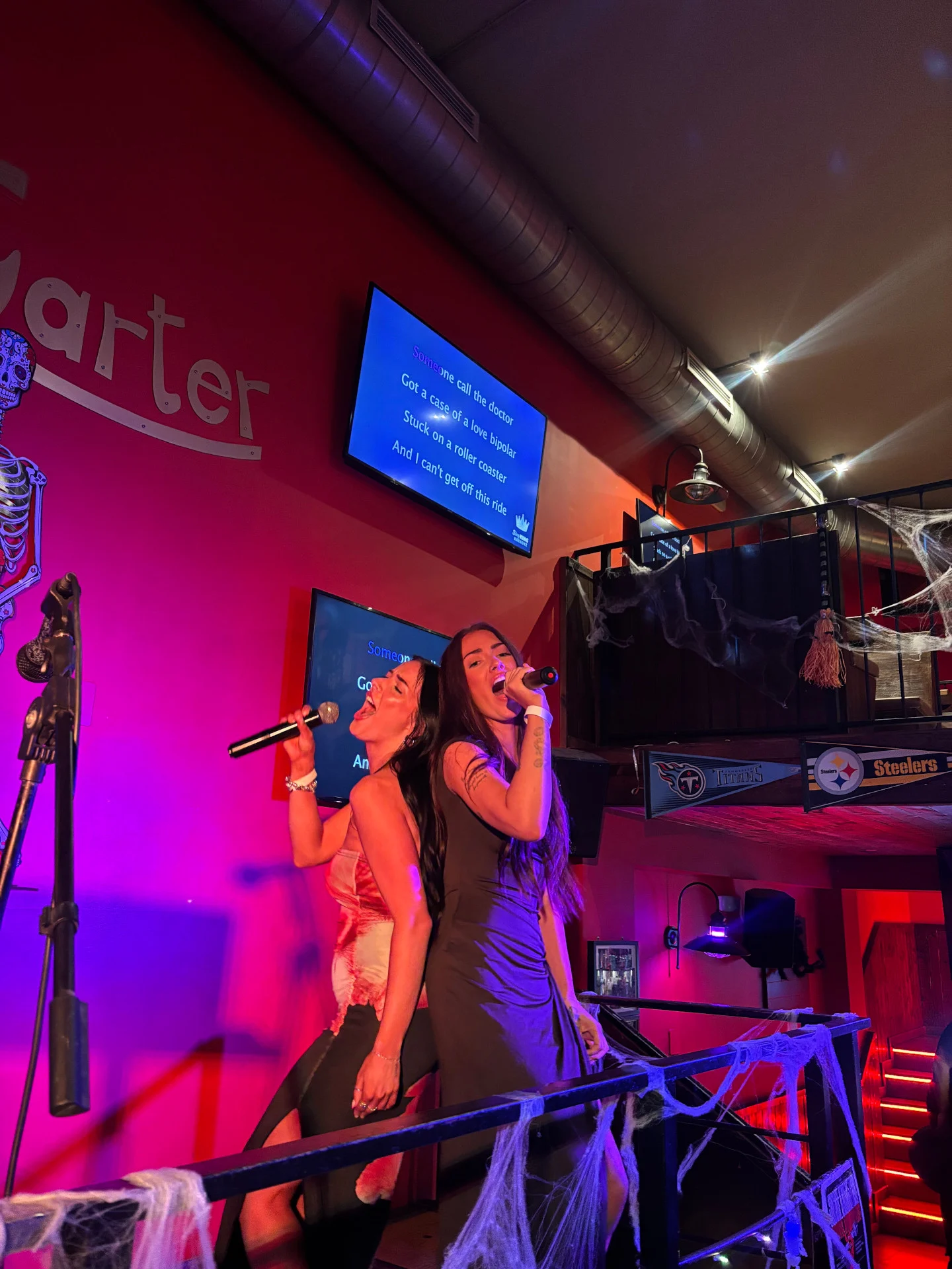 Girls singing on stage at a Barcelona bar, leaning back-to-back with mics in hand as the crowd vibes with the karaoke energy.